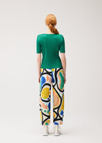 A person with long red hair in a braid wears the PLEATS PLEASE ISSEY MIYAKE ROLL green short-sleeved top and colorful print semi-wide pants, standing facing away against a plain light background.
