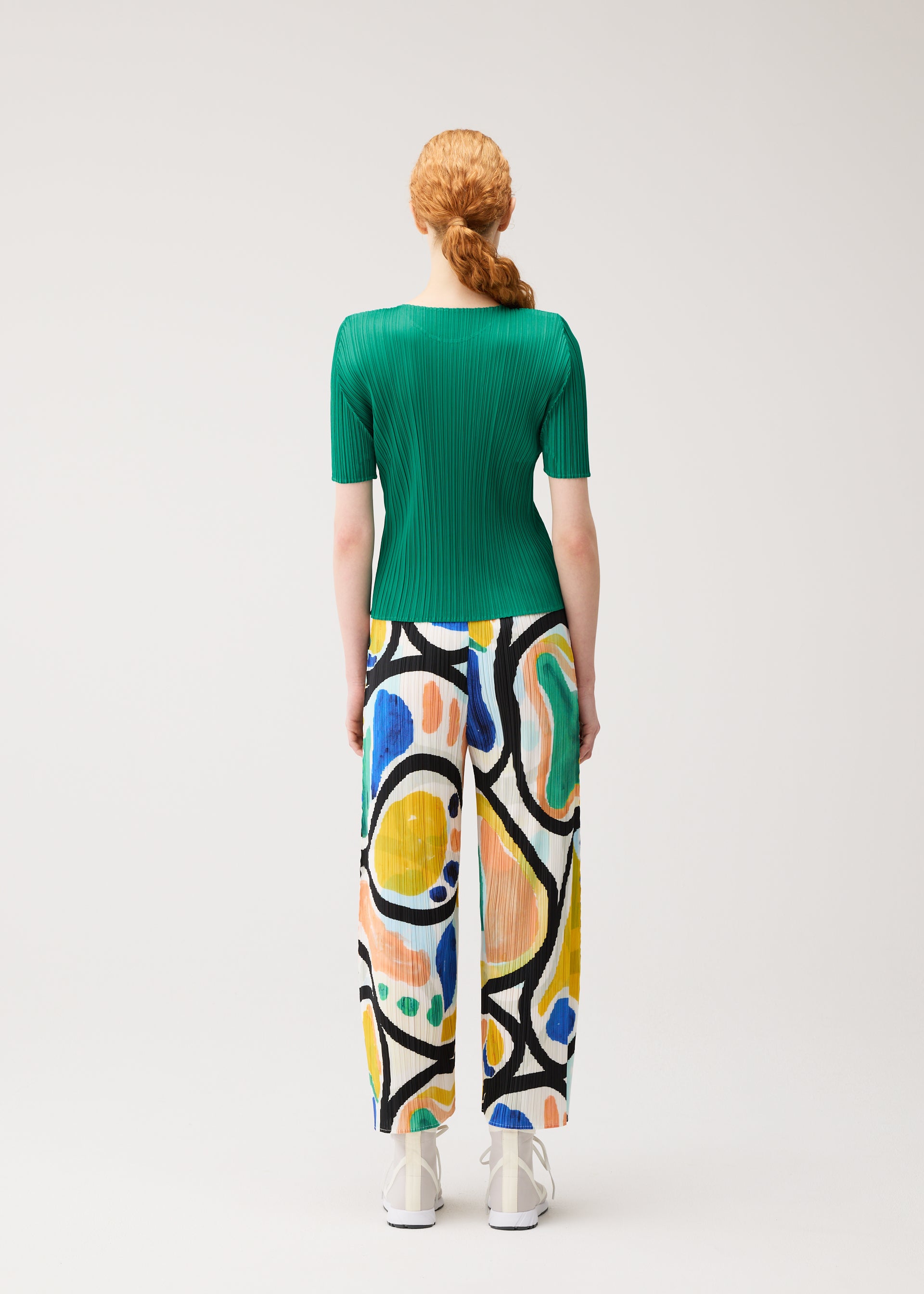 Zoomed view A person with long red hair in a braid wears the PLEATS PLEASE ISSEY MIYAKE ROLL green short-sleeved top and colorful print semi-wide pants, standing facing away against a plain light background.