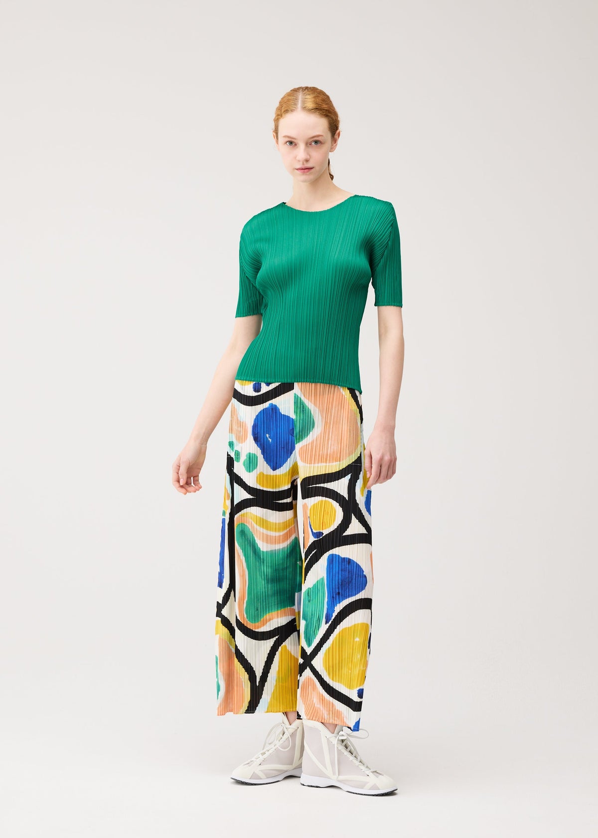 A woman stands against a plain background, wearing the ROLL ribbed green short-sleeve top and abstract print semi-wide pants by PLEATS PLEASE ISSEY MIYAKE, paired with white sneakers. view 7 of 7