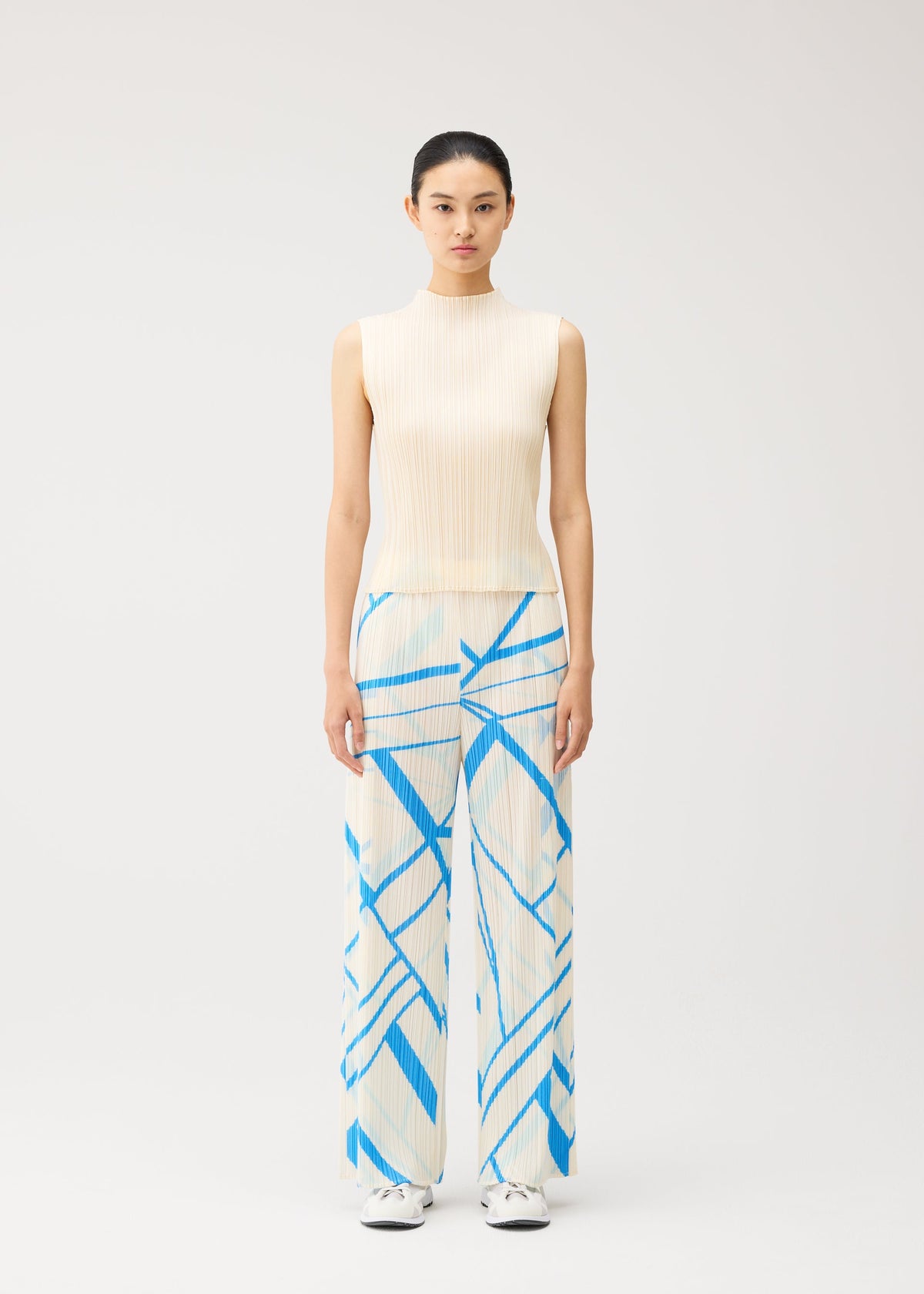 A woman stands facing forward, wearing the FOLD LINE sleeveless cream top and wide-leg trousers with a blue geometric pattern by PLEATS PLEASE ISSEY MIYAKE, featuring a minimalist design. Her hair is tied back, and she is wearing white shoes against a plain, light-colored background. view 5 of 8
