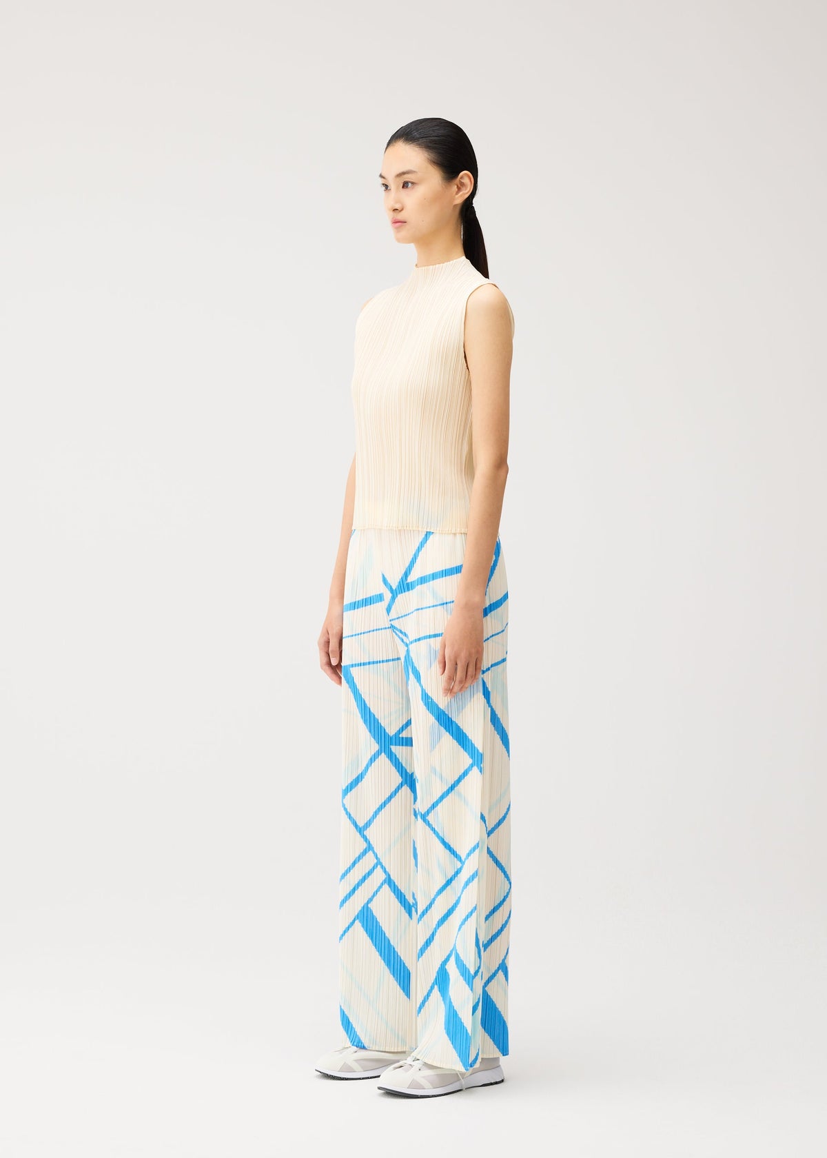 A woman stands sideways against a plain light background, dressed in the FOLD LINE set from PLEATS PLEASE ISSEY MIYAKE—a sleeveless beige top and wide, straight pants adorned with blue geometric patterns and a subtle diagonal wrapping effect—finished with white shoes for a perfect minimalist look. view 6 of 8