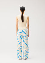 A person with long dark hair in a low ponytail is shown from behind, standing against a plain light background, dressed in the FOLD LINE ensemble by PLEATS PLEASE ISSEY MIYAKE—a sleeveless cream knit top and wide-leg cream pants adorned with minimalist blue geometric stripes.