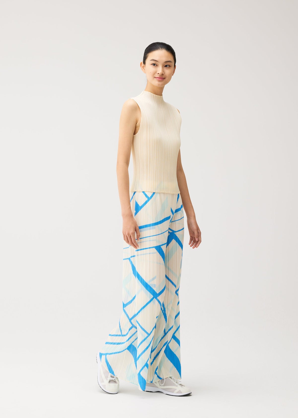 A woman stands wearing the FOLD LINE sleeveless cream top and wide-leg straight pants by PLEATS PLEASE ISSEY MIYAKE, showcasing a minimalist design with blue geometric patterns. Her hair is tied back, she smiles subtly, and she pairs the outfit with white sneakers against a plain light background. view 8 of 8