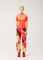 A woman stands facing forward wearing the SURPRISE! short-sleeve top and wide-leg pants from PLEATS PLEASE ISSEY MIYAKE, featuring a bright orange hue and a colorful, uniquely designed pattern, paired with orange lace-up shoes against a plain light background.