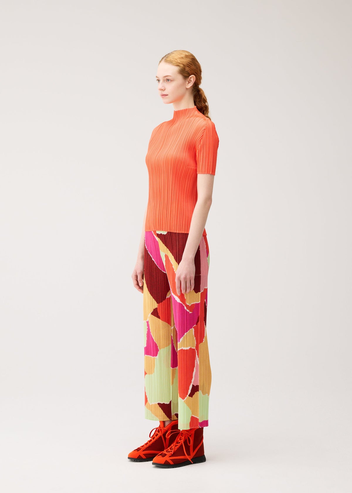 A woman stands in profile against a plain light background, wearing the “SURPRISE!” ribbed top and thin, straight colorful patterned pants from PLEATS PLEASE ISSEY MIYAKE, paired with red high-top sneakers. view 5 of 7