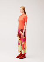 A woman stands in profile against a plain light background, wearing the “SURPRISE!” ribbed top and thin, straight colorful patterned pants from PLEATS PLEASE ISSEY MIYAKE, paired with red high-top sneakers.