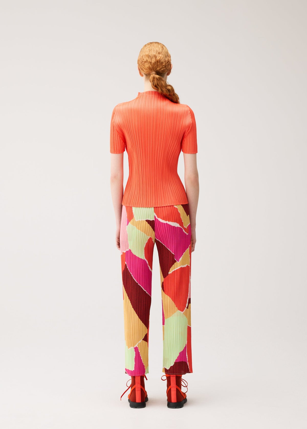 A person with long, red hair styled in a low ponytail is seen from behind, standing against a plain light background and wearing the SURPRISE! set by PLEATS PLEASE ISSEY MIYAKE—a ribbed, short-sleeve orange top paired with thin straight pants featuring a colorful pattern and unique design—and orange shoes. view 6 of 7