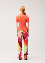 A person with long, red hair styled in a low ponytail is seen from behind, standing against a plain light background and wearing the SURPRISE! set by PLEATS PLEASE ISSEY MIYAKE—a ribbed, short-sleeve orange top paired with thin straight pants featuring a colorful pattern and unique design—and orange shoes.