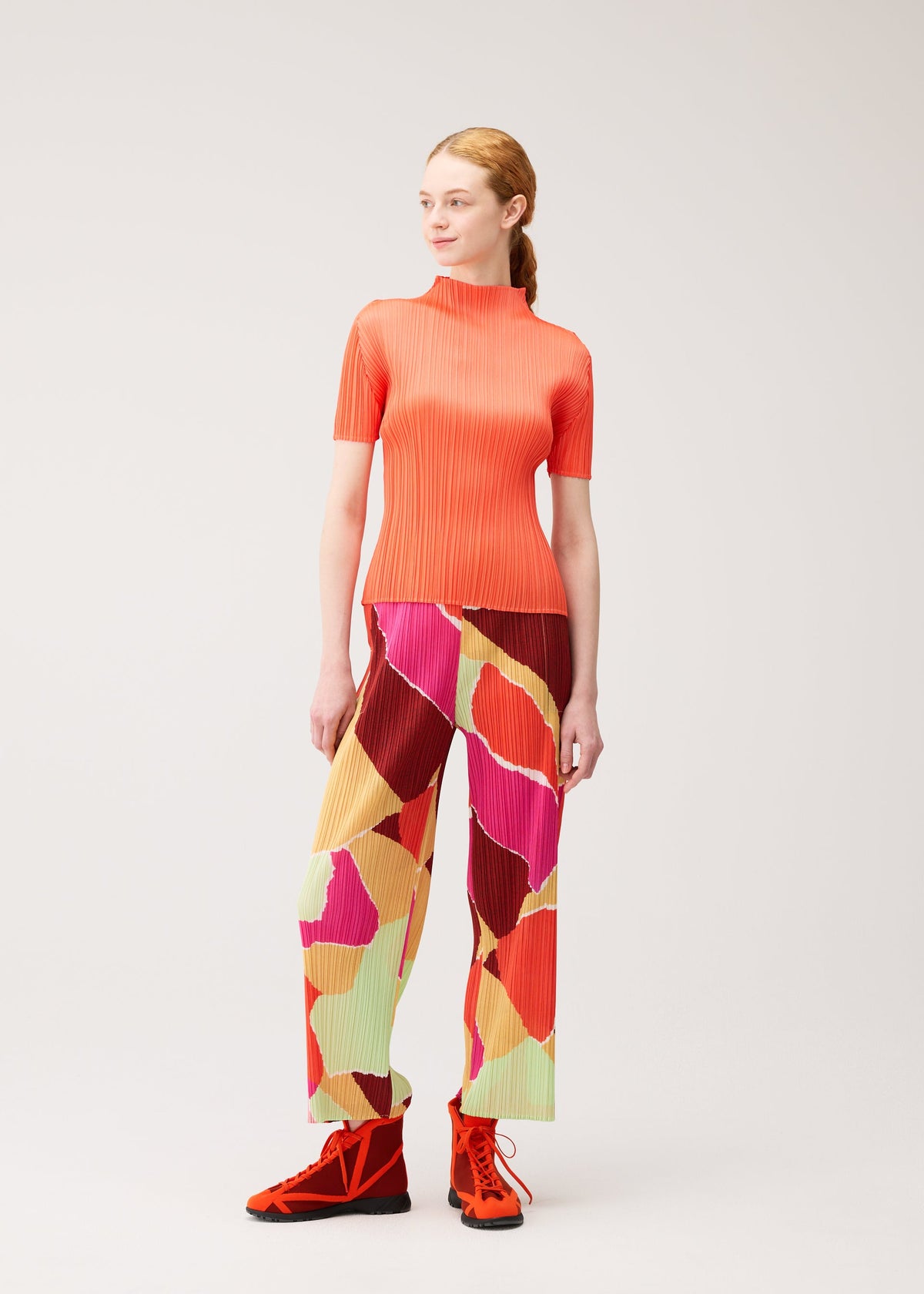 A woman stands against a plain background wearing the SURPRISE! pleated top and thin, straight pants with a colorful pattern and unique design from PLEATS PLEASE ISSEY MIYAKE, paired with red shoes. She looks slightly to the side with her hair tied back. view 7 of 7