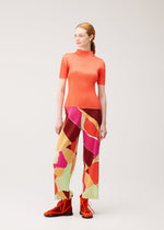 A woman stands against a plain background wearing the SURPRISE! pleated top and thin, straight pants with a colorful pattern and unique design from PLEATS PLEASE ISSEY MIYAKE, paired with red shoes. She looks slightly to the side with her hair tied back.