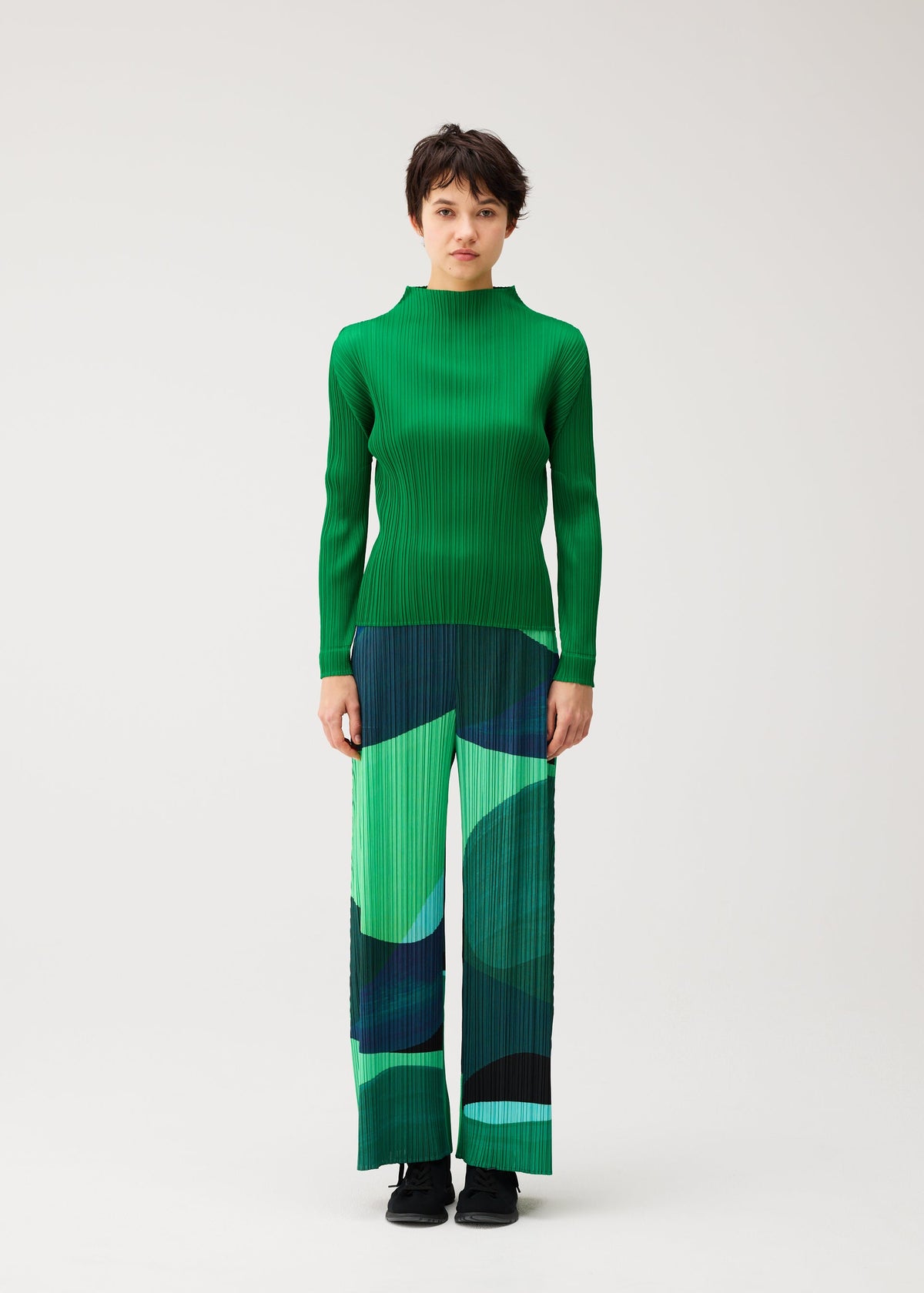 Zoomed view A person stands facing forward in the ORBIT wide pleated pants by PLEATS PLEASE ISSEY MIYAKE—green, blue, and black abstract patterns—with a green long-sleeve top against a plain light background.