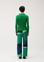 Person stands with back to camera, wearing a green long-sleeve top and ORBIT wide-leg pleated pants by PLEATS PLEASE ISSEY MIYAKE, featuring abstract green, blue, and black overlapping shapes on a light background.