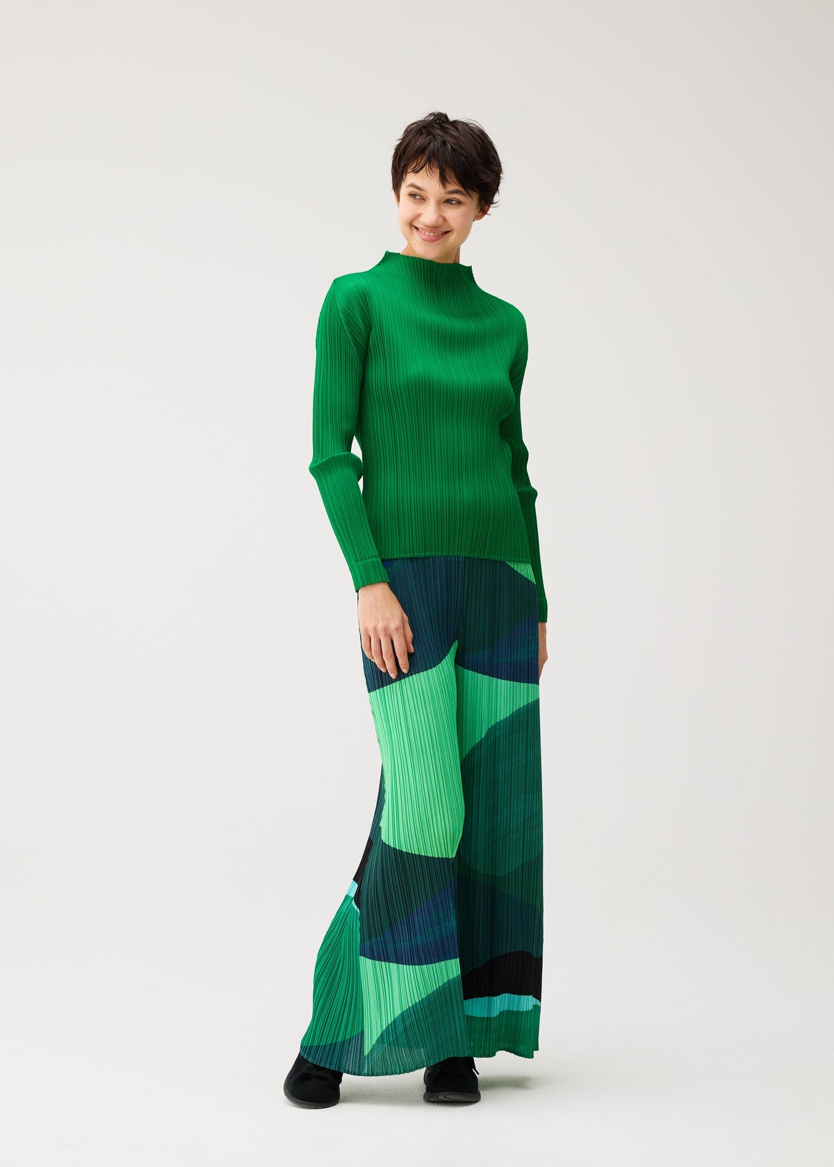 Zoomed view A person models the ORBIT skirt by PLEATS PLEASE ISSEY MIYAKE, featuring overlapping green and dark blue shapes, paired with a green long-sleeve top against a plain light background.