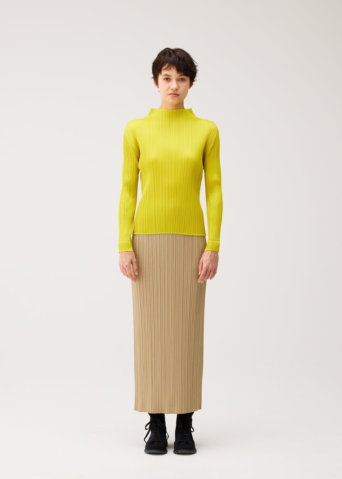 A person stands facing forward in the MONTHLY COLORS : OCTOBER top by PLEATS PLEASE ISSEY MIYAKE, a bright yellow long-sleeve shirt, paired with a beige straight skirt and black boots against a plain white background. view 7 of 10