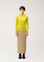 A person stands facing forward in the MONTHLY COLORS : OCTOBER top by PLEATS PLEASE ISSEY MIYAKE, a bright yellow long-sleeve shirt, paired with a beige straight skirt and black boots against a plain white background.