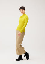 A person models the MONTHLY COLORS : OCTOBER yellow ribbed turtleneck and beige straight skirt with pockets by PLEATS PLEASE ISSEY MIYAKE, posing playfully in black boots against a plain white background.