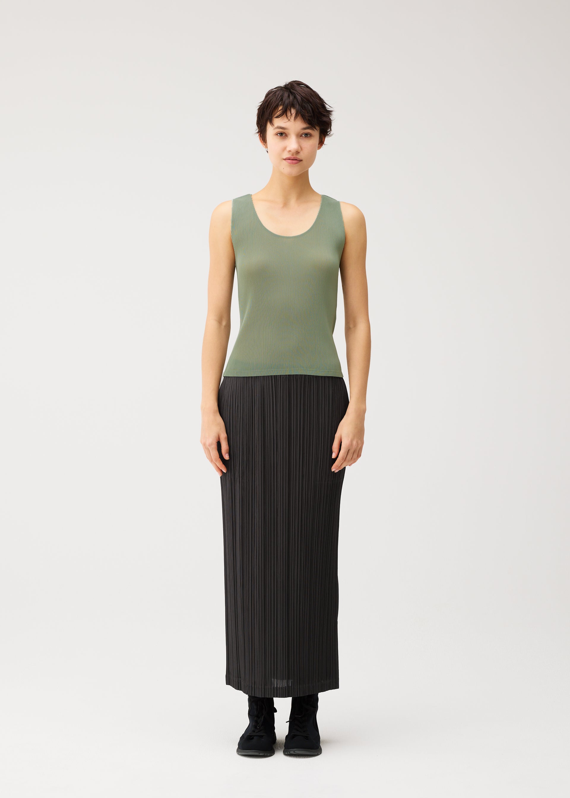Zoomed view A person stands facing forward, dressed in a sleeveless green top and the PLEATS PLEASE ISSEY MIYAKE THICKER BOTTOMS 2—a long black straight skirt with side pockets crafted from thick fabric—paired with black shoes, set against a plain white background.