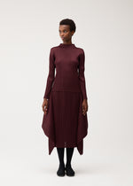 A woman stands against a plain light background, wearing FLUTTER—a long-sleeved, burgundy dress by PLEATS PLEASE ISSEY MIYAKE. The dress features a distinctive pleated skirt with an uneven hem and is paired with black tights and black shoes. She gazes slightly to the side, her arms relaxed at her sides.