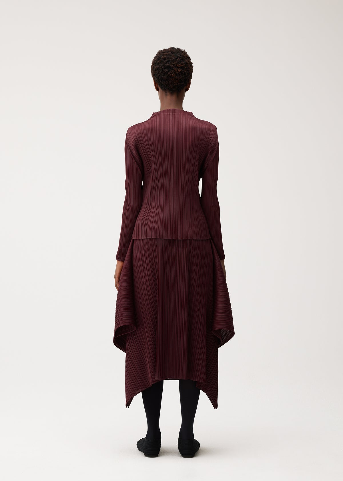 Zoomed view A person stands facing away against a plain light background, wearing the FLUTTER long-sleeved pleated dress by PLEATS PLEASE ISSEY MIYAKE in burgundy. The dress features a distinctive quadrilateral skirt with an asymmetrical hem, paired with black tights and black shoes.