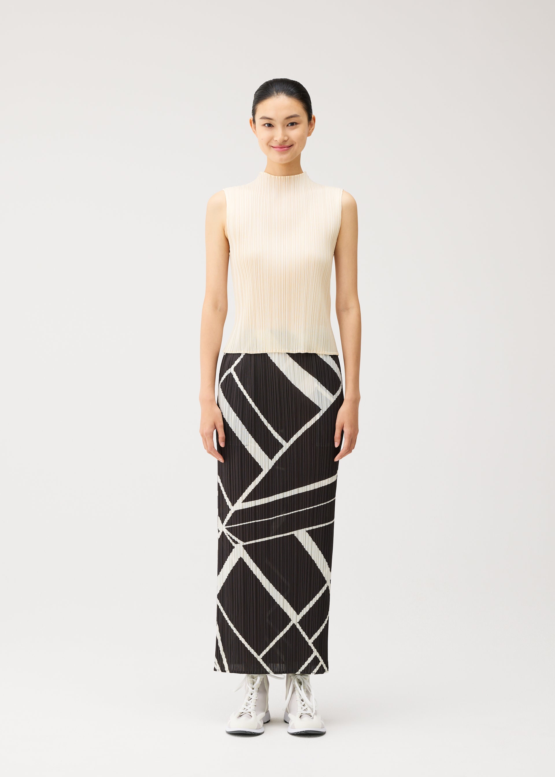 Zoomed view A woman stands facing forward, smiling, dressed in the FOLD LINE skirt by PLEATS PLEASE ISSEY MIYAKE—a long, straight black skirt with bold white geometric lines in a minimalist design—paired with a sleeveless cream top and white lace-up shoes against a plain light background.
