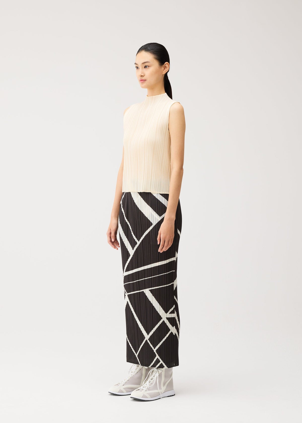 A woman stands in profile against a plain light background, wearing the FOLD LINE long black straight skirt with white geometric patterns from PLEATS PLEASE ISSEY MIYAKE. She pairs it with a sleeveless beige top and white lace-up ankle boots, creating a refined, modern look with her minimalist ensemble. view 5 of 7