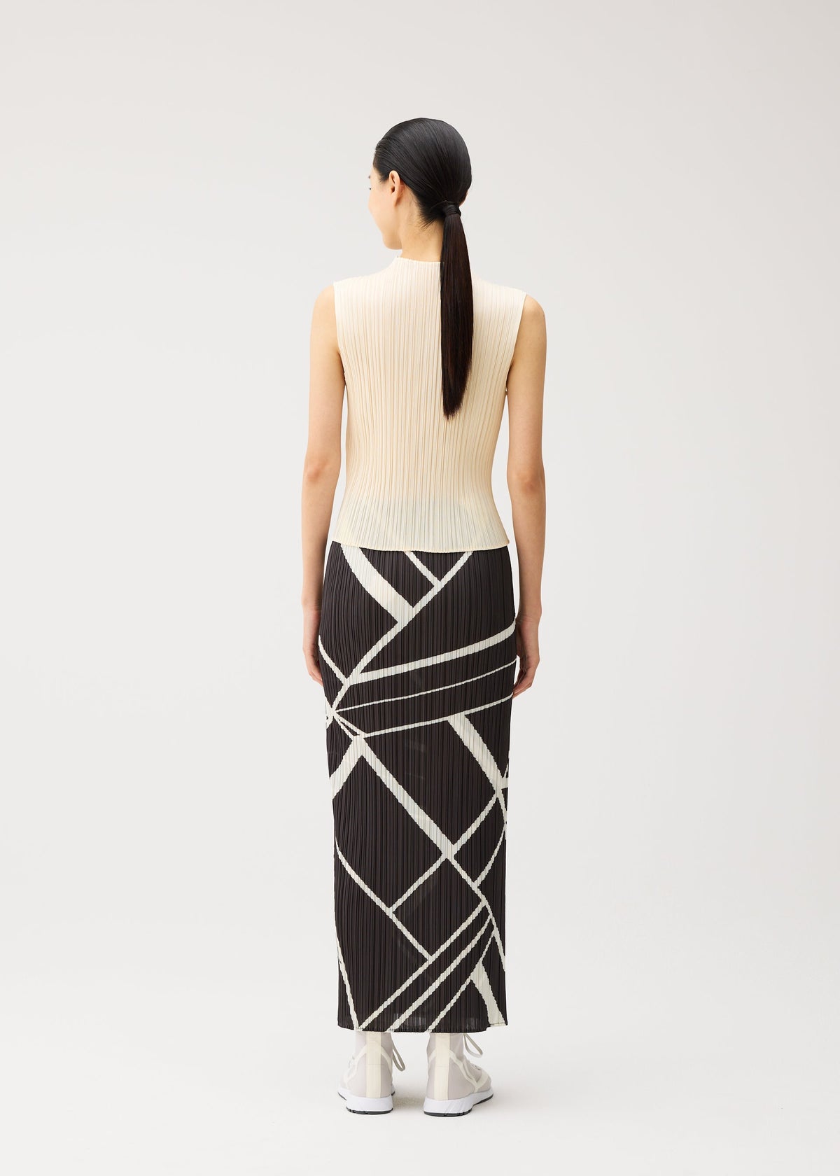 A woman with long dark hair in a ponytail models the FOLD LINE ensemble by PLEATS PLEASE ISSEY MIYAKE—a sleeveless cream top paired with a long, straight black skirt featuring white geometric lines—standing facing away against a light background. The minimalist design of the outfit highlights clean lines and understated elegance. view 6 of 7