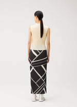 A woman with long dark hair in a ponytail models the FOLD LINE ensemble by PLEATS PLEASE ISSEY MIYAKE—a sleeveless cream top paired with a long, straight black skirt featuring white geometric lines—standing facing away against a light background. The minimalist design of the outfit highlights clean lines and understated elegance.