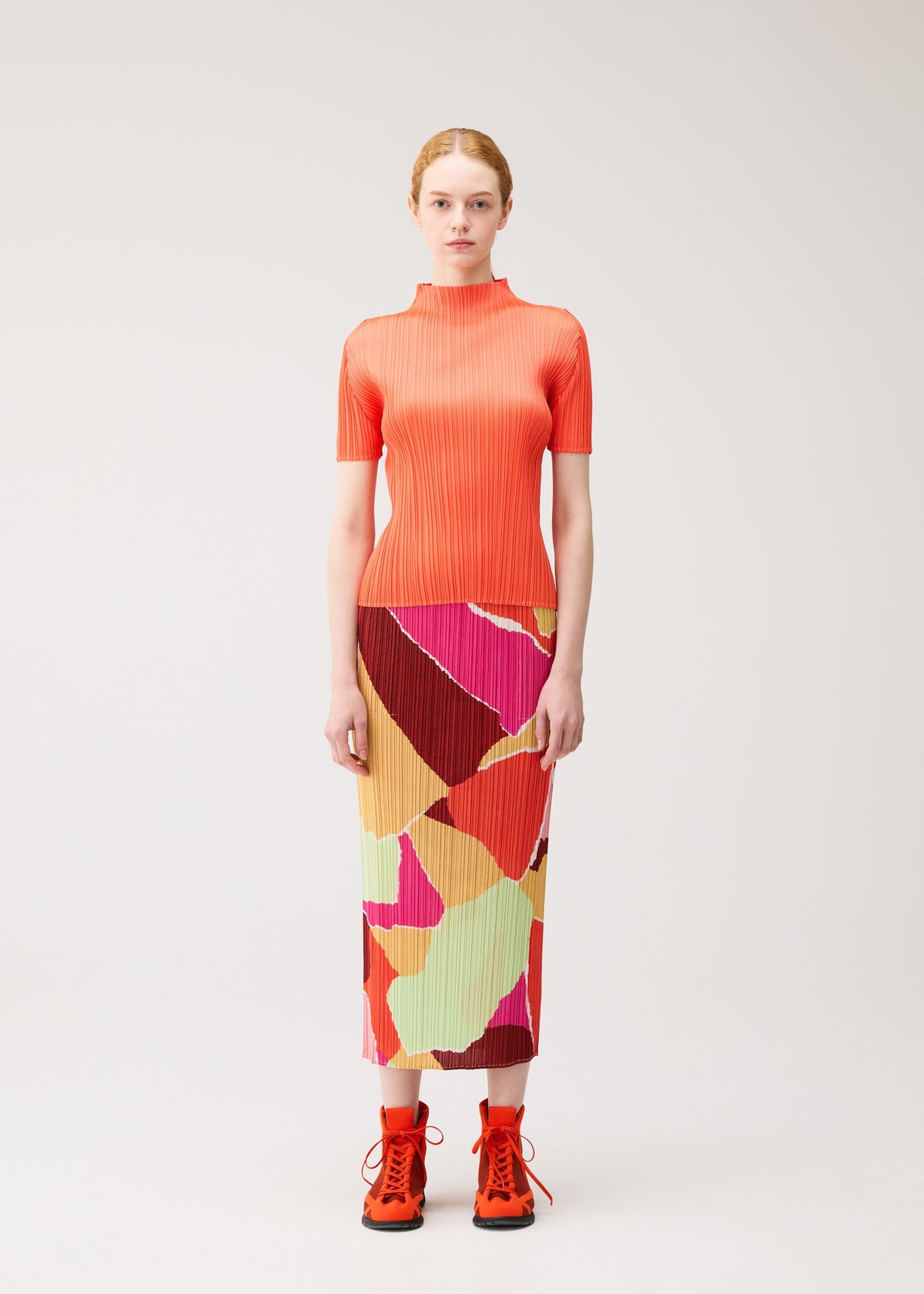 A woman stands facing forward, wearing the vibrant ribbed “SURPRISE!” top and matching straight skirt with pockets from PLEATS PLEASE ISSEY MIYAKE, both featuring a colorful pattern of abstract shapes. She completes the look with bright orange lace-up shoes against a plain light background. view 4 of 7