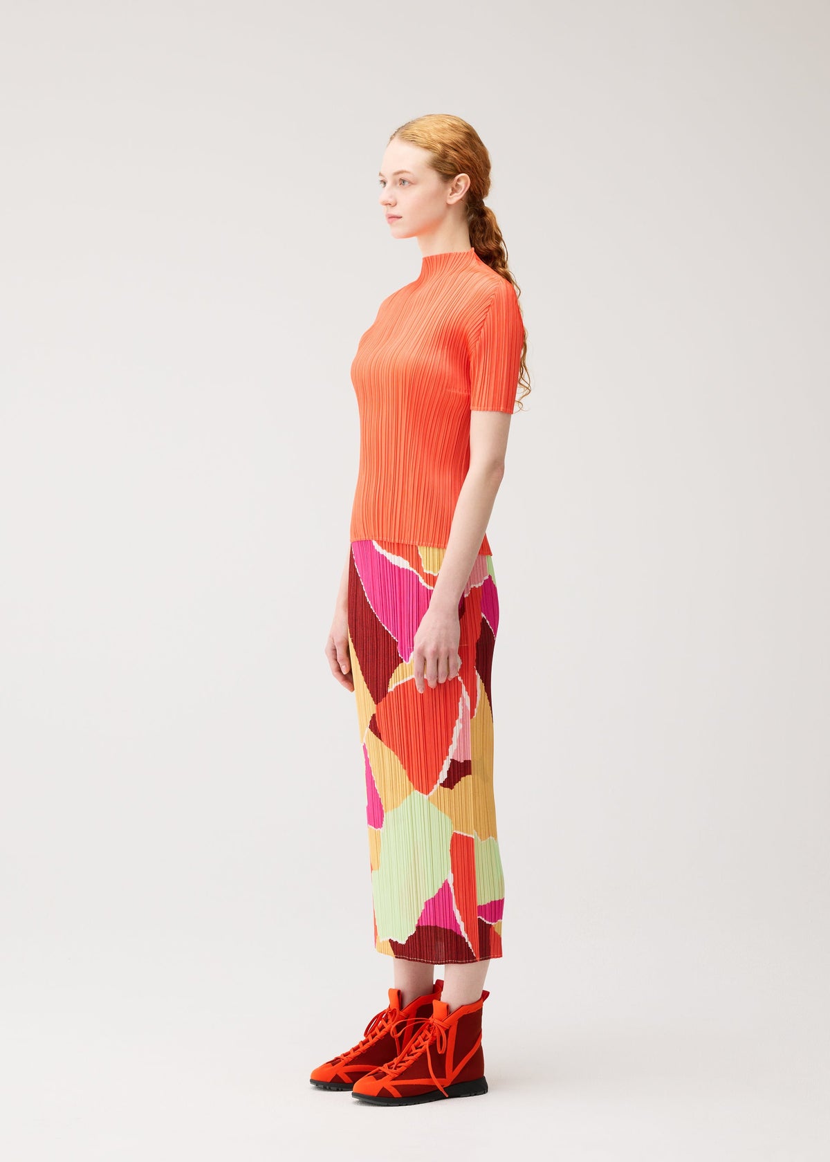 A woman stands sideways against a plain light background, wearing the SURPRISE! pleated top and straight skirt with handy pockets in a colorful pattern by PLEATS PLEASE ISSEY MIYAKE, paired with bright orange lace-up shoes. view 5 of 7