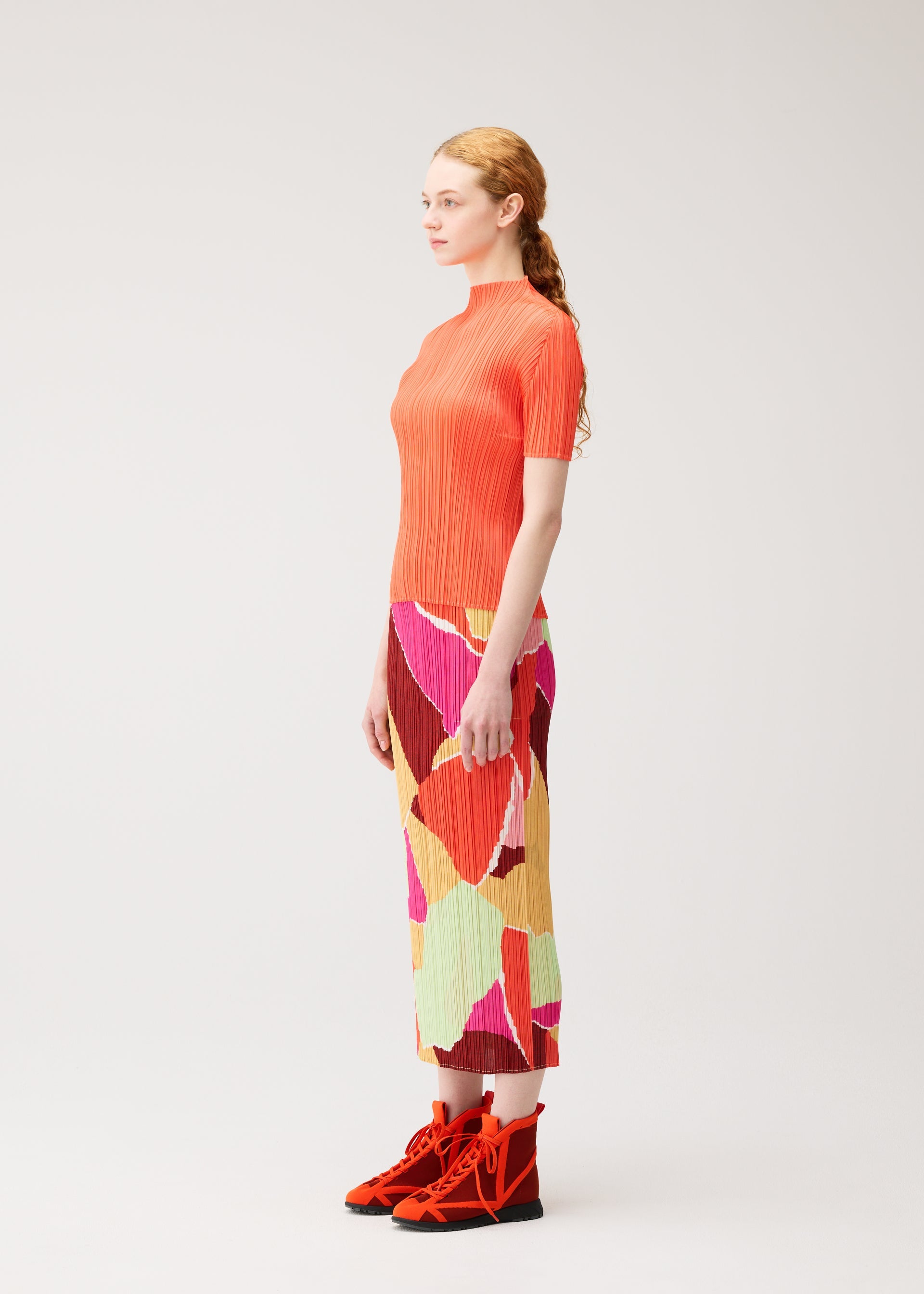 Zoomed view A woman stands sideways against a plain light background, wearing the SURPRISE! pleated top and straight skirt with handy pockets in a colorful pattern by PLEATS PLEASE ISSEY MIYAKE, paired with bright orange lace-up shoes.
