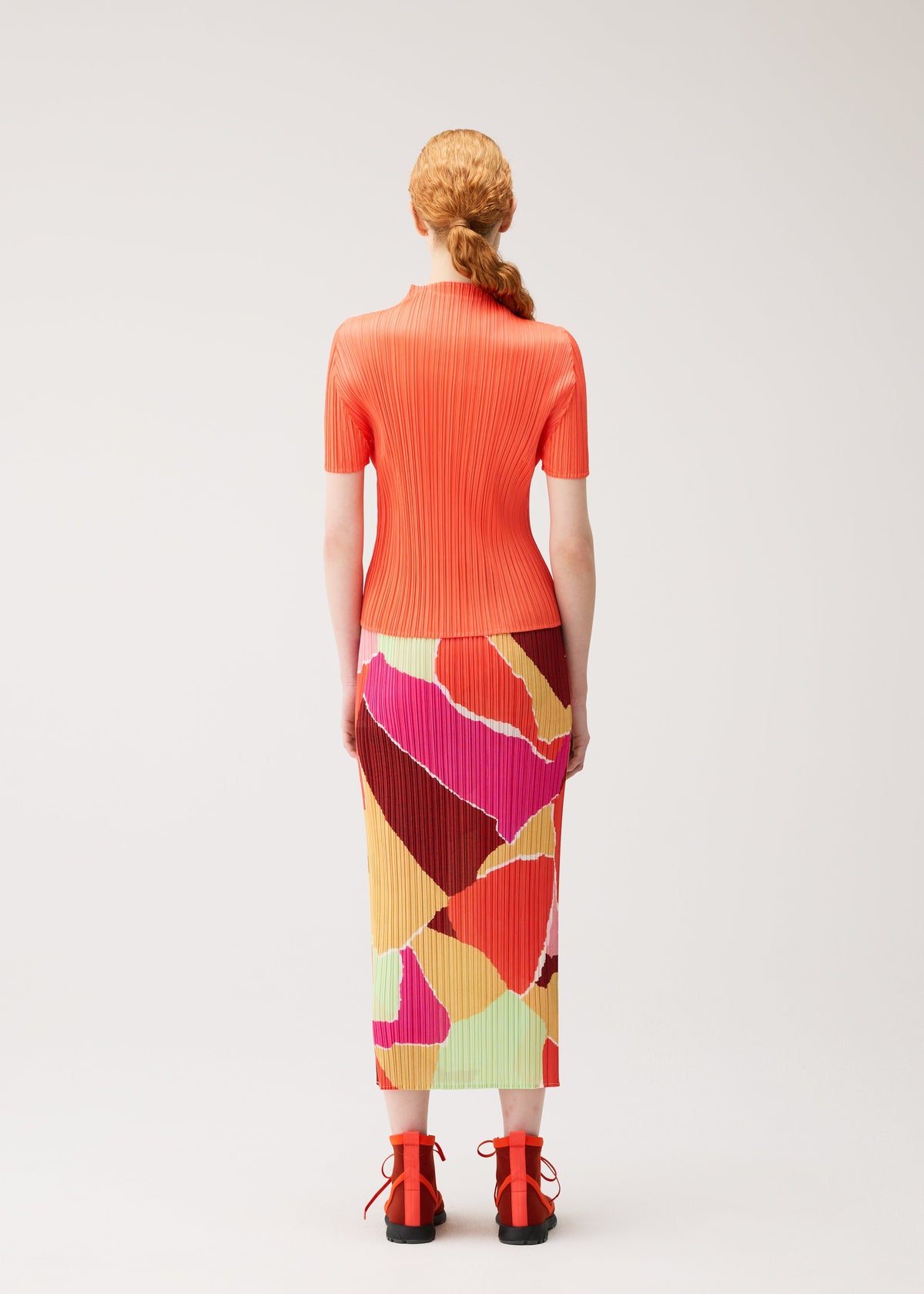 A person with red hair in a low ponytail, wearing the SURPRISE! bright orange ribbed top and a colorful straight skirt with an abstract pattern from PLEATS PLEASE ISSEY MIYAKE, along with red shoes, stands facing away against a plain white background. view 6 of 7