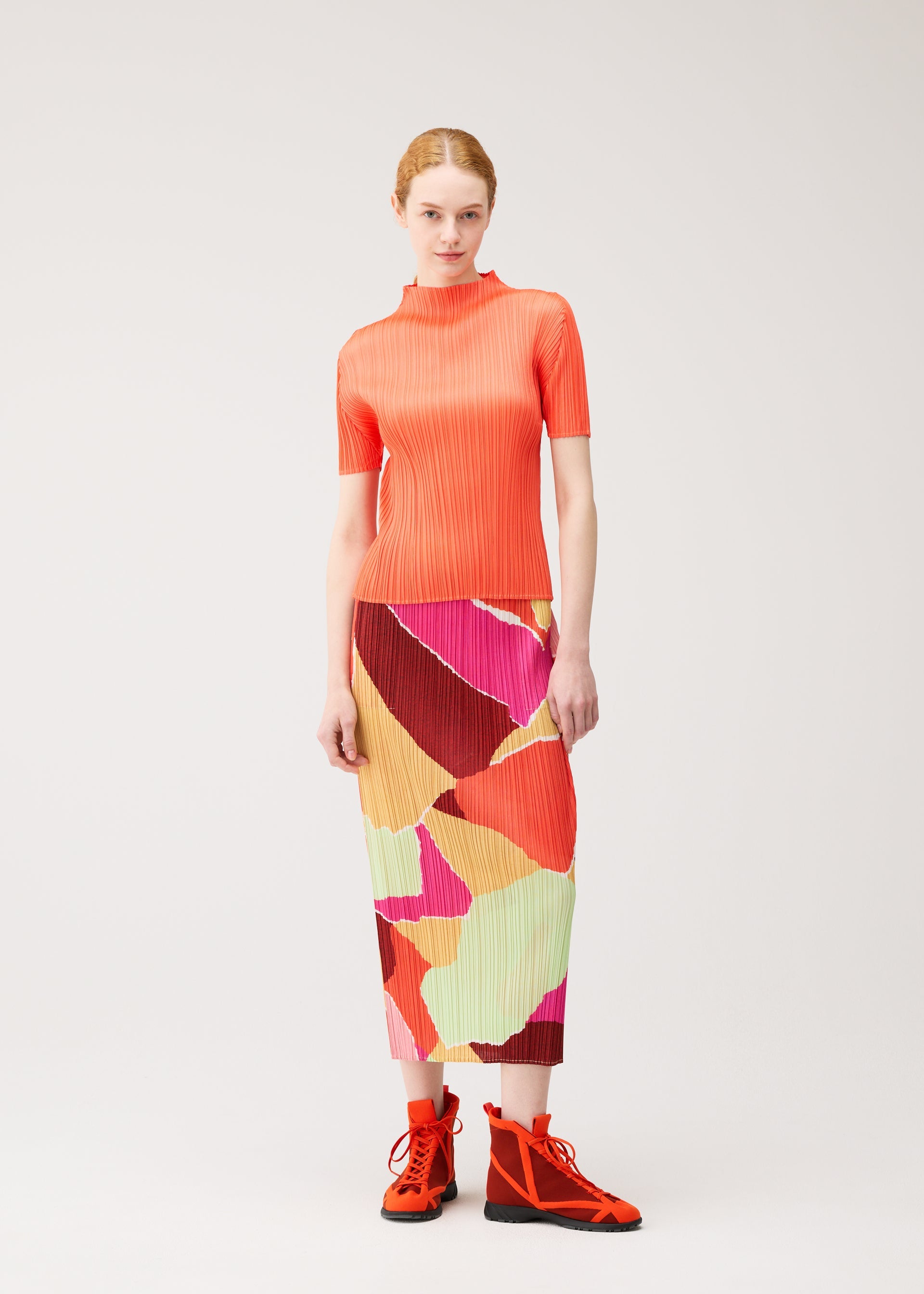 Zoomed view A woman stands against a plain background wearing the SURPRISE! pleated orange top by PLEATS PLEASE ISSEY MIYAKE, a colorful patterned skirt, and bright orange high-top shoes.