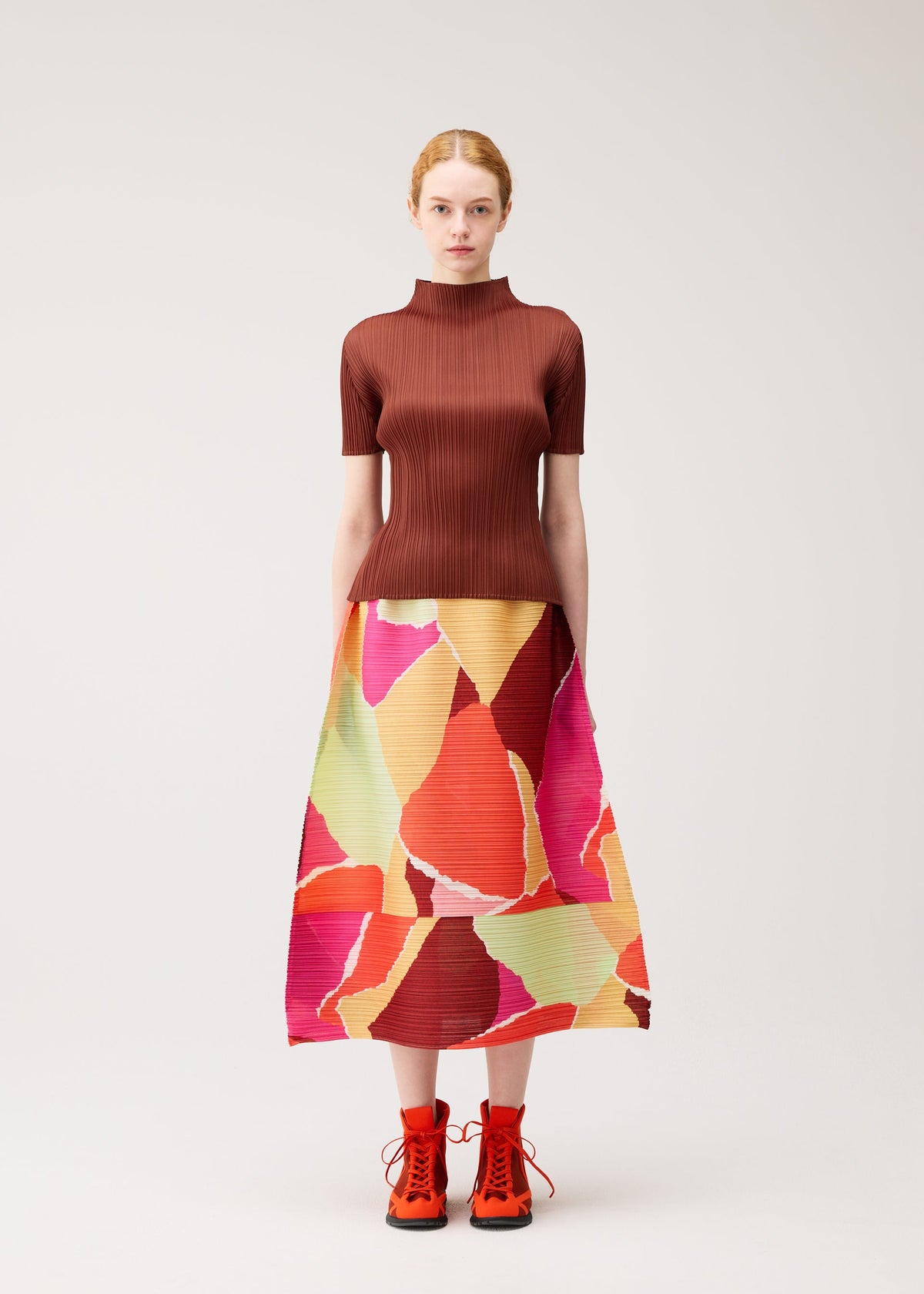 A woman stands against a plain background, wearing the “SURPRISE!” pleated skirt by PLEATS PLEASE ISSEY MIYAKE, featuring a unique pattern in vibrant red, pink, orange, and green abstract designs. She pairs it with a brown ribbed top and bright red lace-up boots. view 4 of 7