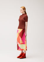 A woman stands sideways against a plain light background, wearing the PLEATS PLEASE ISSEY MIYAKE “SURPRISE!” colorful pleated skirt, paired with a brown ribbed top and red ankle boots.