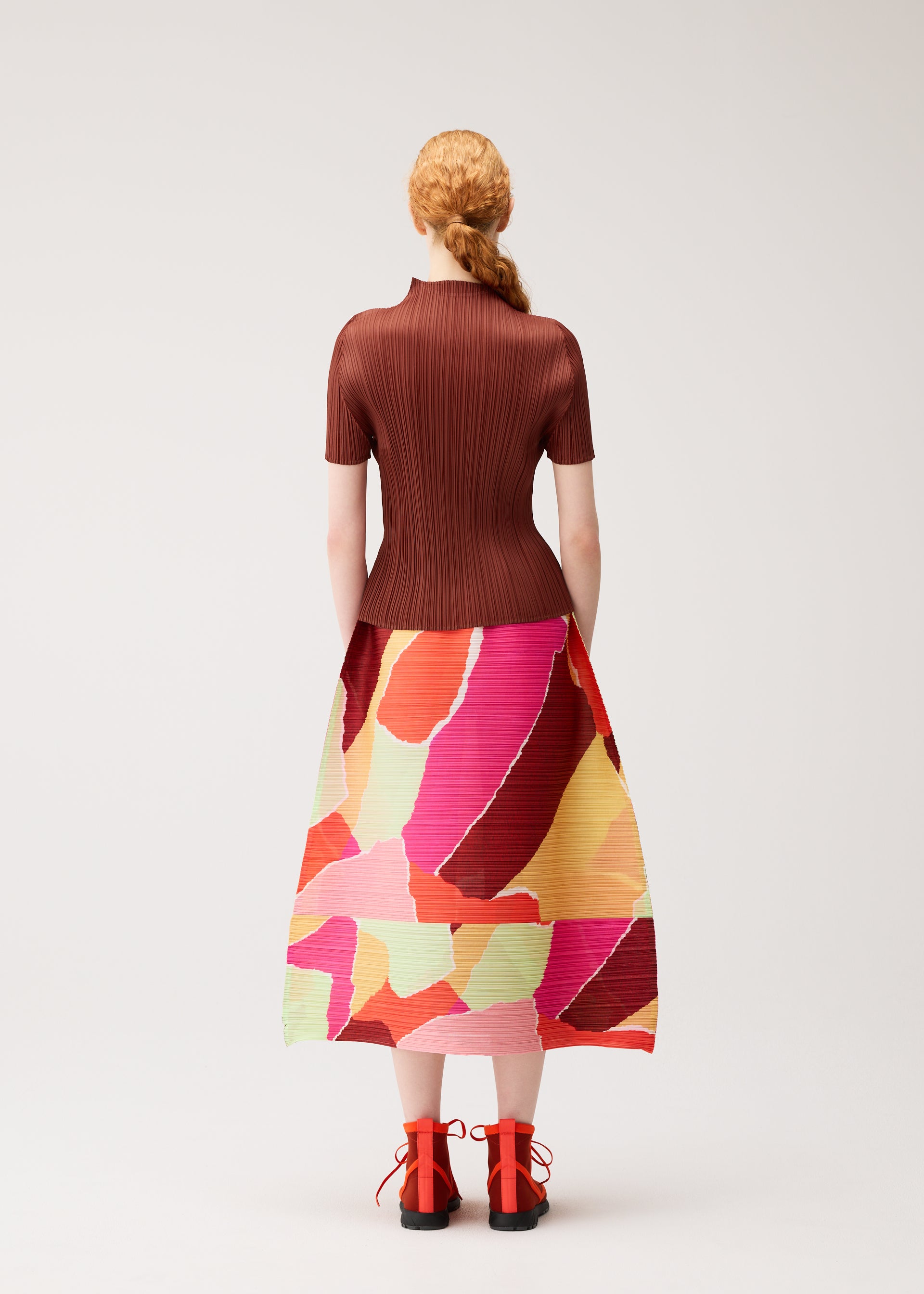 Zoomed view A person with red hair styled in a braid wears the SURPRISE! skirt by PLEATS PLEASE ISSEY MIYAKE, featuring a colorful and unique pattern, along with a ribbed brown top and red shoes. They are standing facing away against a plain light background.