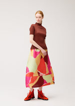 A woman stands wearing the “SURPRISE!” colorful pleated skirt by PLEATS PLEASE ISSEY MIYAKE, featuring a unique pattern of red, pink, and green abstract shapes, paired with a brown short-sleeve top and bright red lace-up shoes against a plain light background.