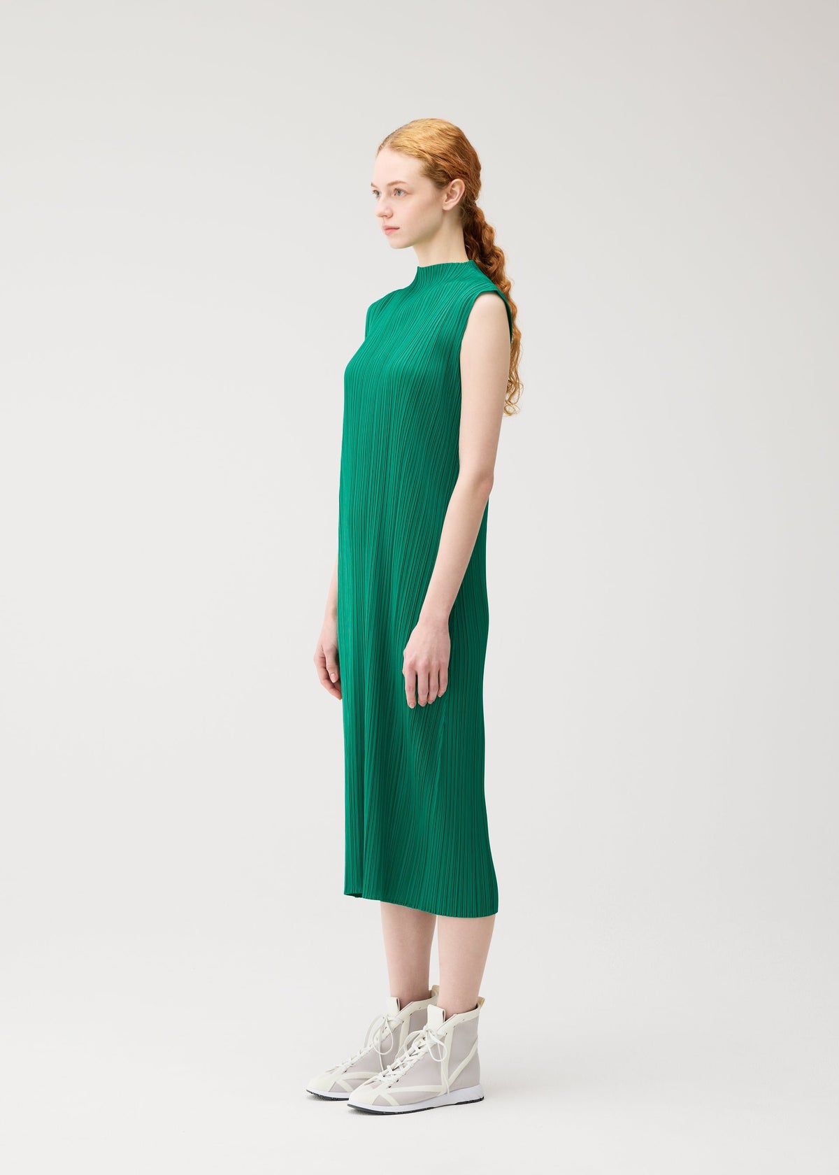 A woman with long, red braided hair stands in profile against a plain light background, wearing the PLEATS PLEASE ISSEY MIYAKE MONTHLY COLORS : JULY sleeveless green high-neck dress and white high-top shoes. view 8 of 10