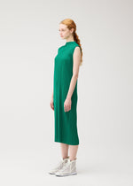A woman with long, red braided hair stands in profile against a plain light background, wearing the PLEATS PLEASE ISSEY MIYAKE MONTHLY COLORS : JULY sleeveless green high-neck dress and white high-top shoes.