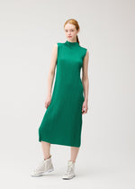 A woman with light skin and red hair tied back models the MONTHLY COLORS : JULY sleeveless green pleated knee-length high-neck dress by PLEATS PLEASE ISSEY MIYAKE and white high-top sneakers, against a plain light background.