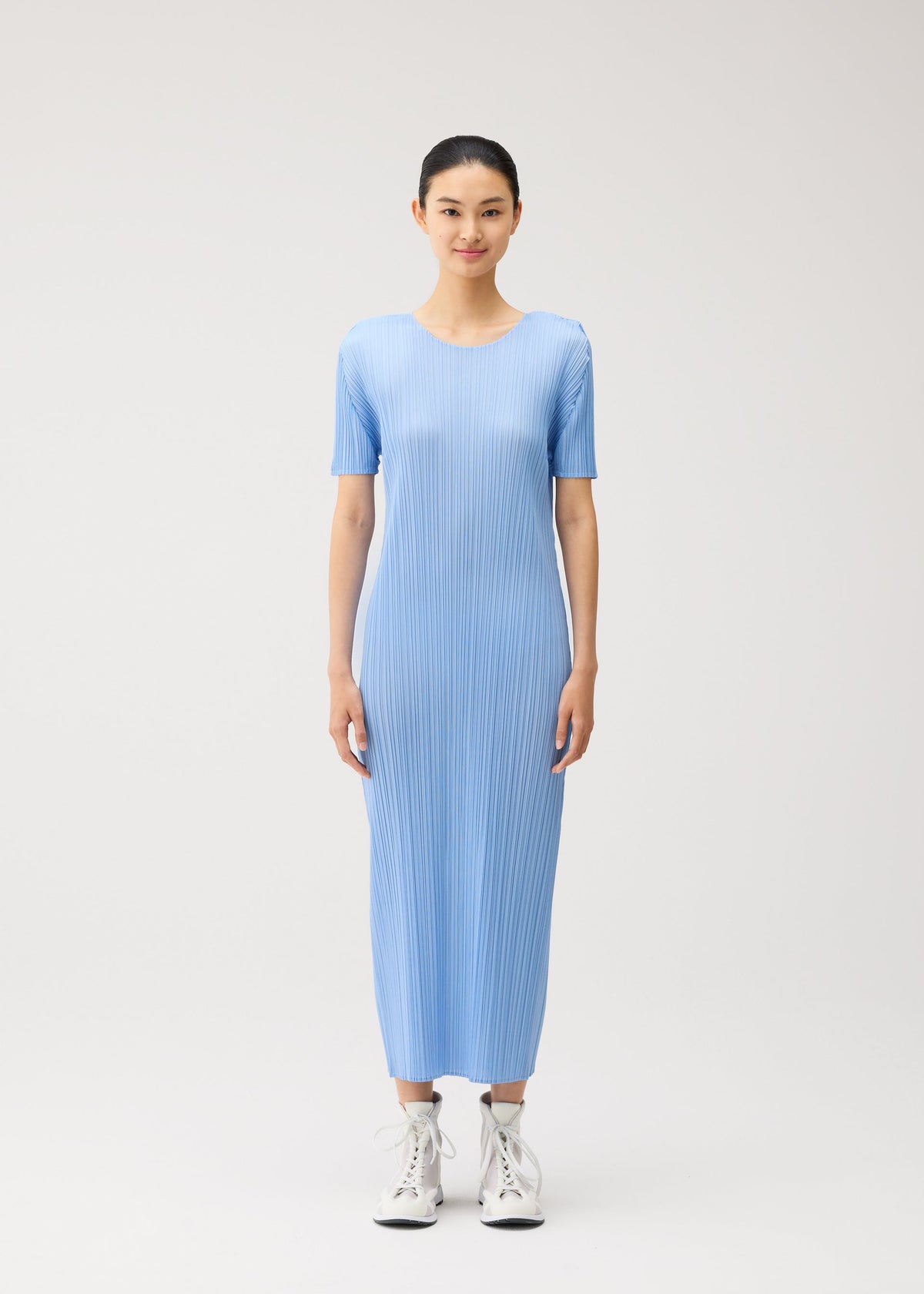 A woman stands facing forward, wearing the MONTHLY COLORS : AUGUST light blue, short-sleeved pleated midi dress by PLEATS PLEASE ISSEY MIYAKE with white high-top sneakers against a plain, light background. view 7 of 10