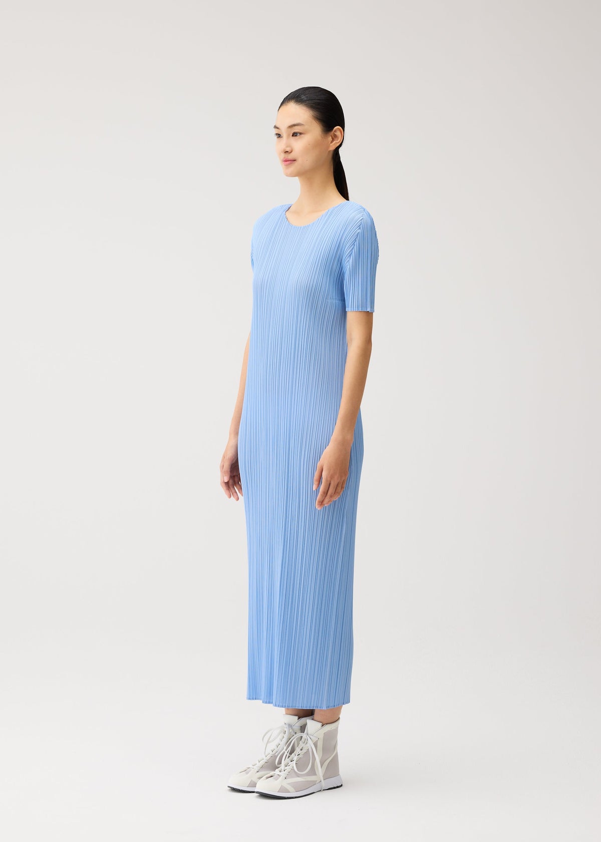 A woman stands against a plain, light background wearing the MONTHLY COLORS : AUGUST dress from PLEATS PLEASE ISSEY MIYAKE—a ribbed, light blue, ankle-length summer dress with short sleeves and a round neckline—paired with white high-top sneakers. view 8 of 10