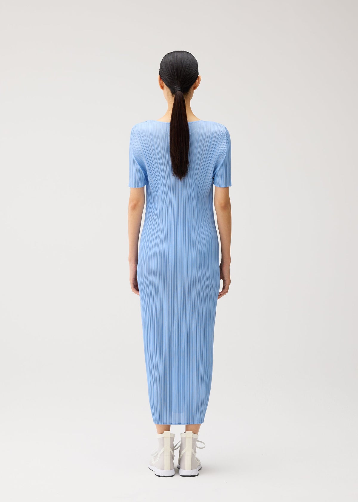 A person with long dark hair in a ponytail is seen from behind, wearing the MONTHLY COLORS : AUGUST blue ribbed short-sleeved summer dress by PLEATS PLEASE ISSEY MIYAKE, featuring a round neckline and ankle-length hem, paired with white shoes against a plain light background. view 9 of 10