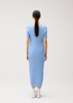 A person with long dark hair in a ponytail is seen from behind, wearing the MONTHLY COLORS : AUGUST blue ribbed short-sleeved summer dress by PLEATS PLEASE ISSEY MIYAKE, featuring a round neckline and ankle-length hem, paired with white shoes against a plain light background.