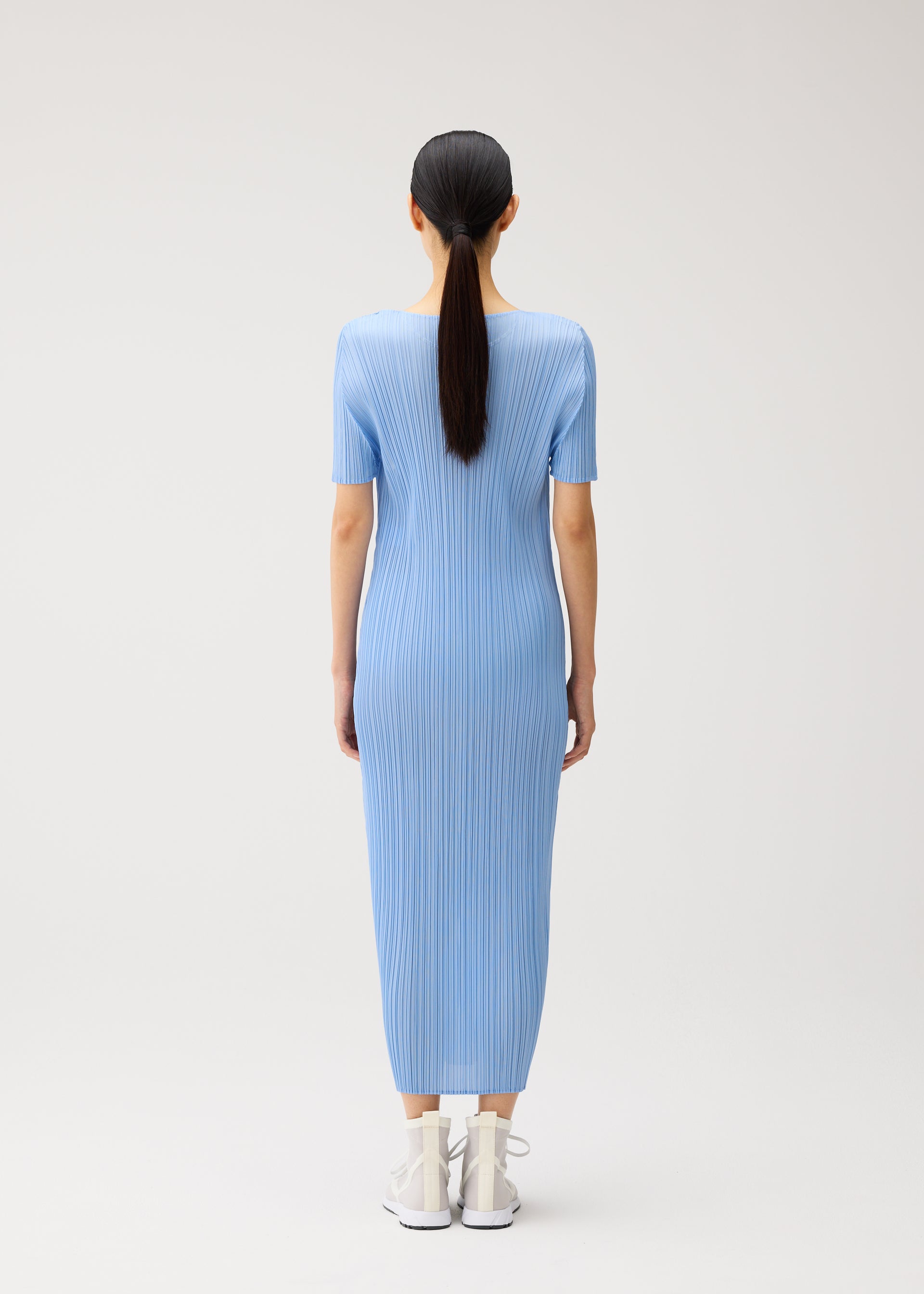 Zoomed view A person with long dark hair in a ponytail is seen from behind, wearing the MONTHLY COLORS : AUGUST blue ribbed short-sleeved summer dress by PLEATS PLEASE ISSEY MIYAKE, featuring a round neckline and ankle-length hem, paired with white shoes against a plain light background.