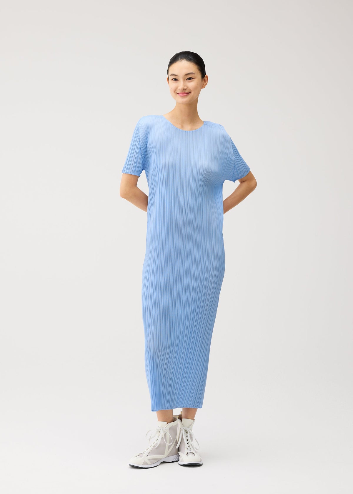 A woman stands against a plain background, smiling with her hands behind her back and wearing white sneakers. She is dressed in the PLEATS PLEASE ISSEY MIYAKE MONTHLY COLORS : AUGUST collection—a light blue, short-sleeved summer dress featuring a round neckline and a pleated skirt. view 10 of 10