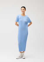 A woman stands against a plain background, smiling with her hands behind her back and wearing white sneakers. She is dressed in the PLEATS PLEASE ISSEY MIYAKE MONTHLY COLORS : AUGUST collection—a light blue, short-sleeved summer dress featuring a round neckline and a pleated skirt.