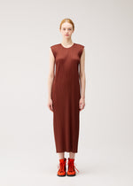 A woman stands facing forward against a plain, light background, wearing the ankle-length, round-neck sleeveless pleated dress from PLEATS PLEASE ISSEY MIYAKE’s MONTHLY COLORS : SEPTEMBER collection in gentle brown tones, paired with bright orange lace-up shoes—a look perfectly suited for the changing seasons.