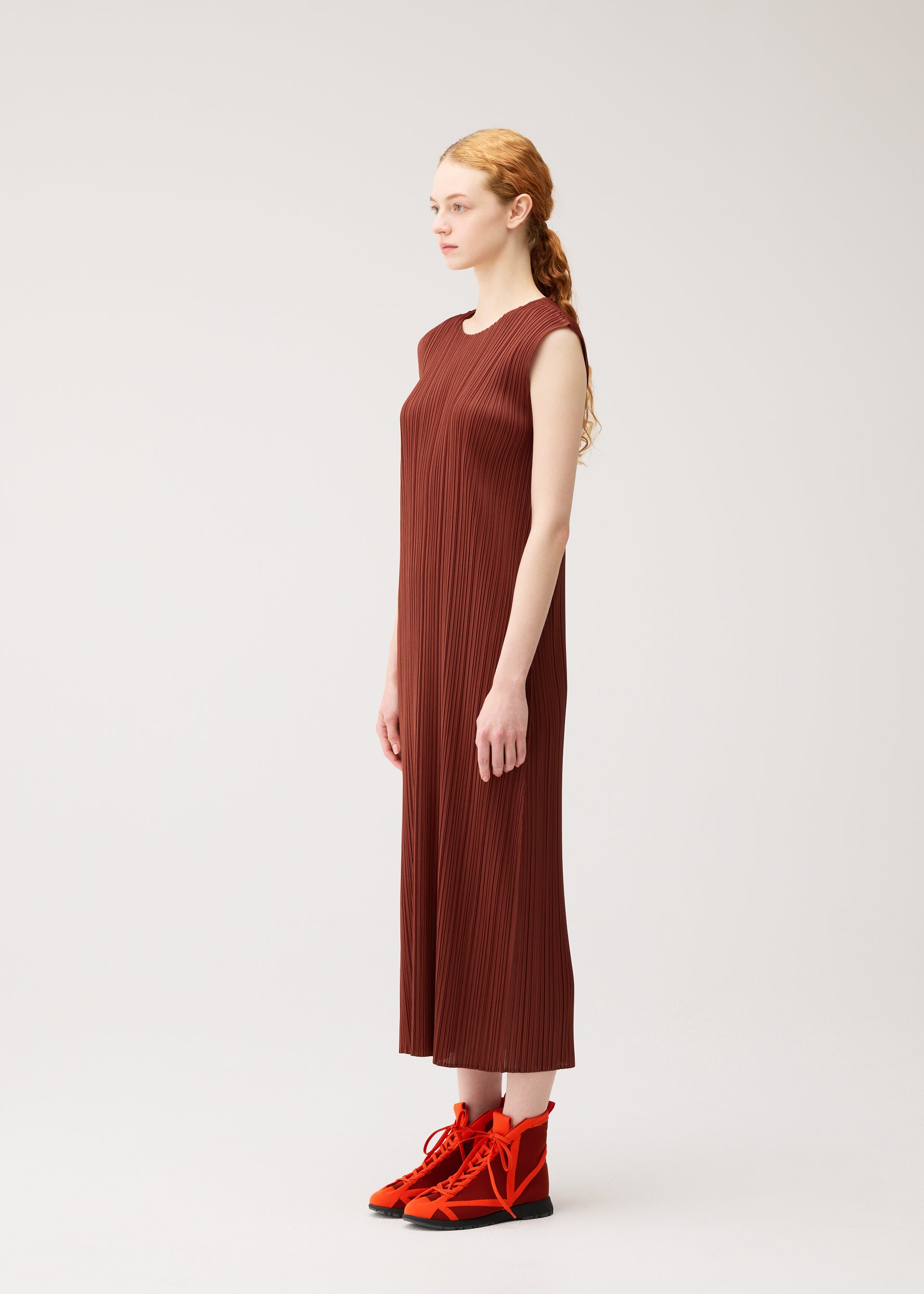 Zoomed view A woman with long blonde hair in a low ponytail stands in profile against a plain light background, wearing the MONTHLY COLORS : SEPTEMBER ankle-length, round-neck sleeveless brown pleated dress by PLEATS PLEASE ISSEY MIYAKE in gentle tones, paired with bright red lace-up boots.