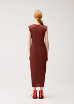 A person with braided red hair wears the MONTHLY COLORS : SEPTEMBER dress from PLEATS PLEASE ISSEY MIYAKE—a round-neck, sleeveless, ankle-length brown pleated dress in gentle tones—paired with red lace-up shoes, standing against a plain white background and facing away from the camera.