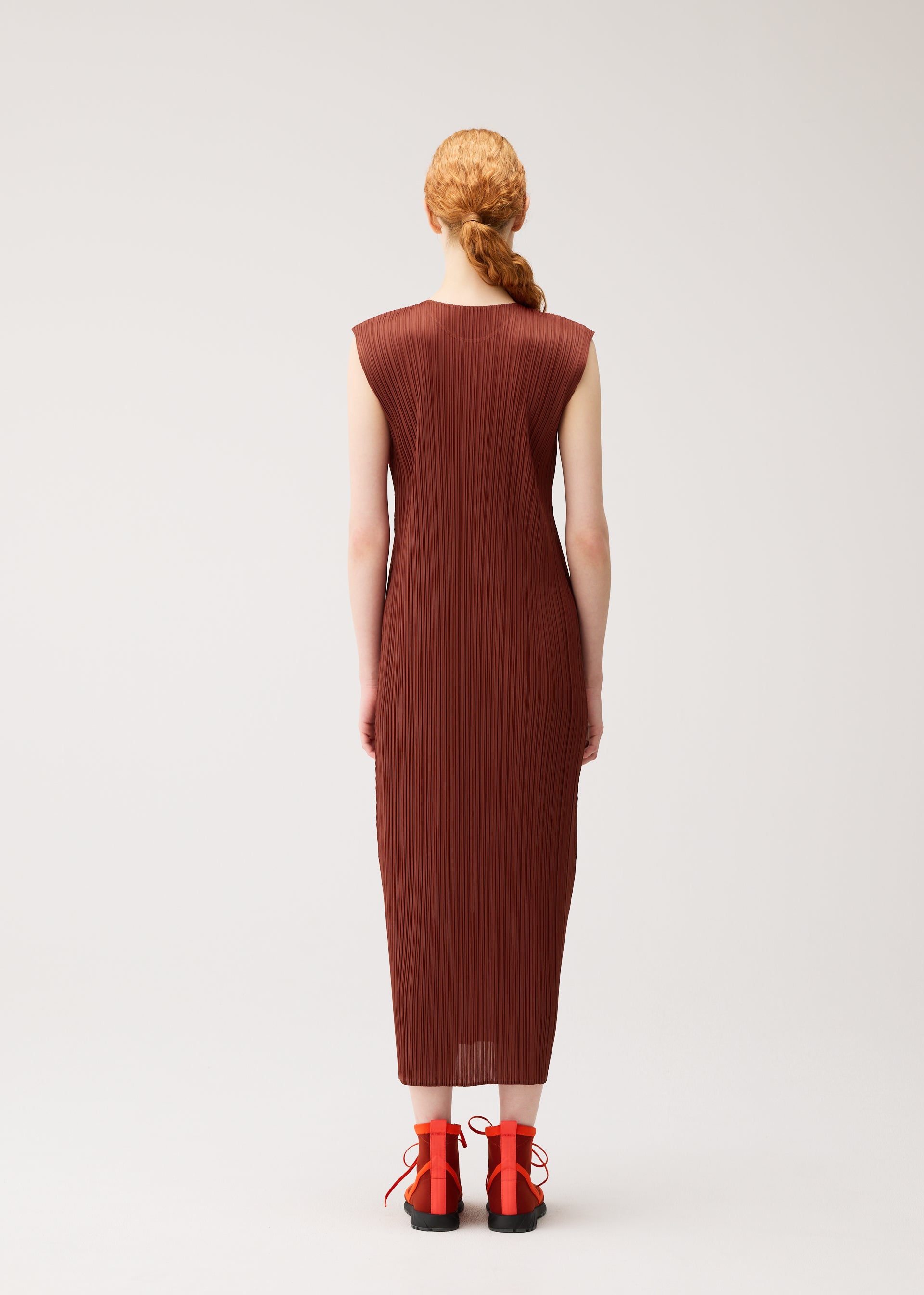Zoomed view A person with braided red hair wears the MONTHLY COLORS : SEPTEMBER dress from PLEATS PLEASE ISSEY MIYAKE—a round-neck, sleeveless, ankle-length brown pleated dress in gentle tones—paired with red lace-up shoes, standing against a plain white background and facing away from the camera.